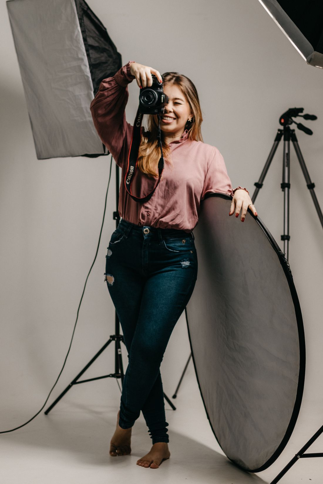 Усміхнена фотографиня з фотоапаратом стоїть біля відбивача й софтбокса у світлій студії